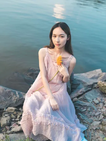 一位穿着粉红色连衣裙的美女 手里拿着一朵花 坐在湖边的一块岩石上。