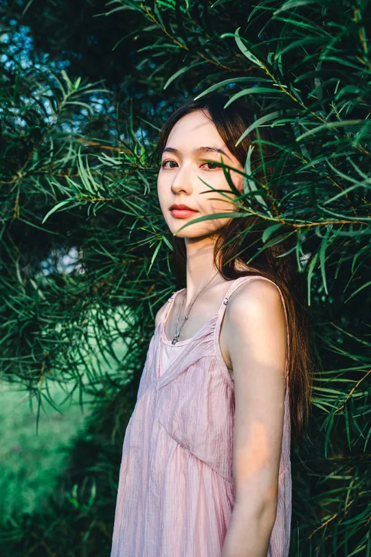 一位穿着粉红色连衣裙的年轻女子站在绿灌木丛和树木前