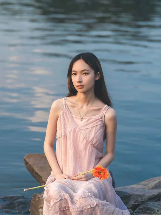 一位穿着粉红色连衣裙的年轻女子坐在湖边的一块岩石上 手里拿着一朵花。