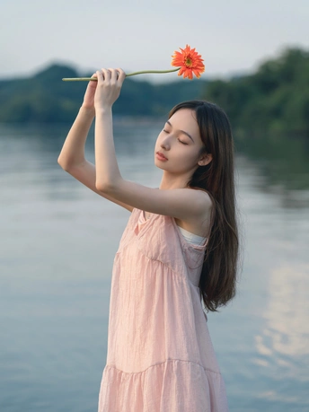 一位穿着粉红色连衣裙的年轻女子手里拿着一朵红花站在水边