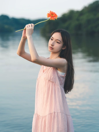 一位穿着粉红色连衣裙的年轻女子手里拿着一朵红花站在水边