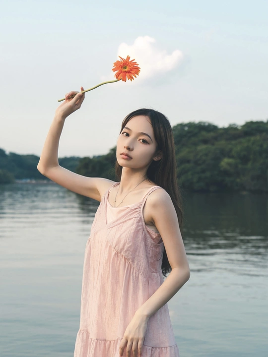 一位穿着粉红色连衣裙的年轻女子手里拿着一朵花站在湖中的水边。