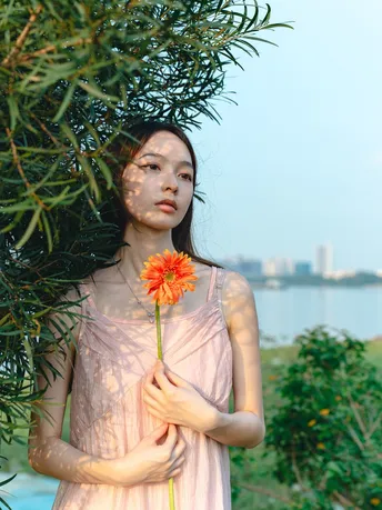 一位穿着粉红色连衣裙的年轻女子手里拿着一朵花 并朝水边望去。