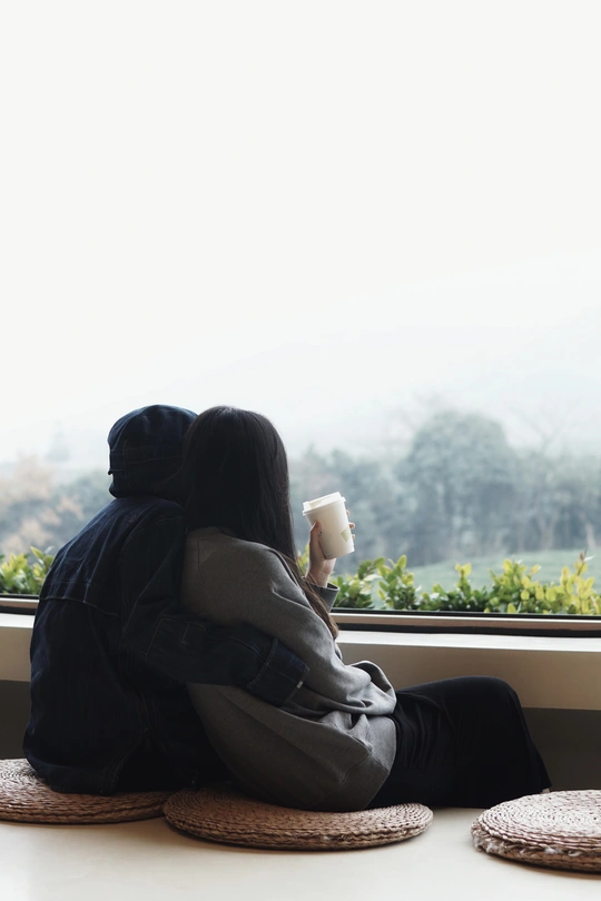 一个年轻女子坐在窗户旁边 手里拿着一杯咖啡 同时看着窗外 而另一个人则坐在窗户上。