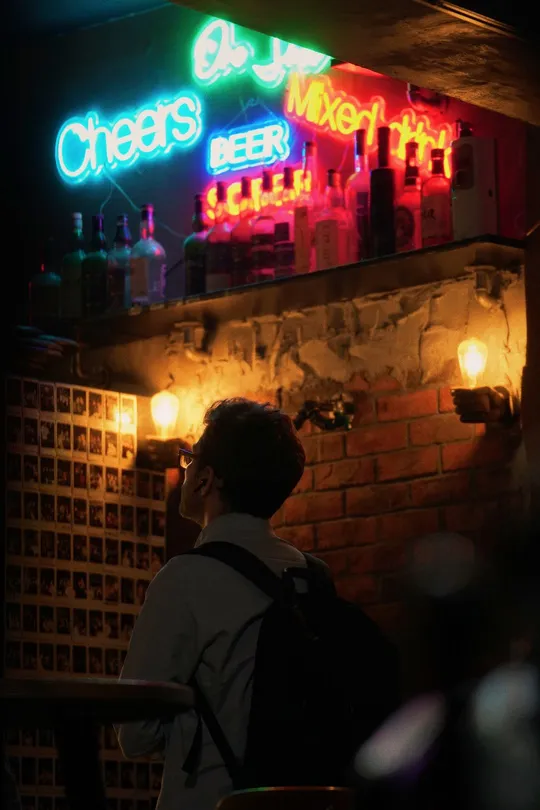 一个男人晚上坐在酒吧里 正在给站在墙上的发光氖灯标志拍照 一个人站在那里。