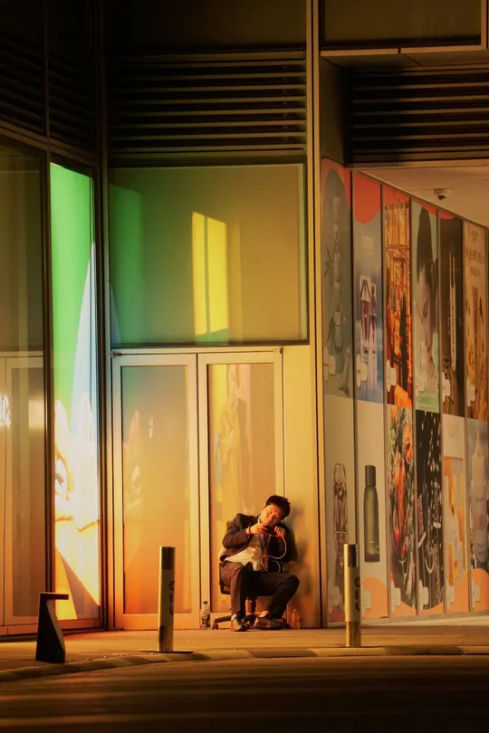 一个坐在长凳上 在人行道上弹吉他的男子 前方有一栋建筑 墙上挂着图片。