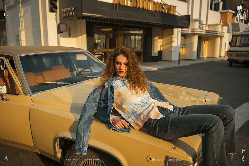 一个穿牛仔夹克的女青年坐在黄色汽车的车盖上在街上