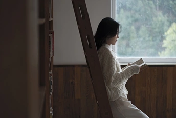 一个年轻女子站在木梯上 正在看一本书 并透过窗户向外看。