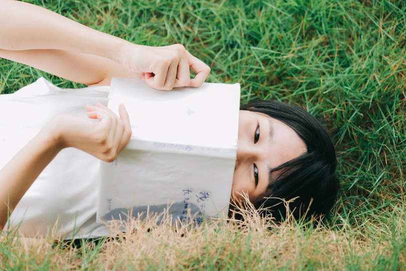 一位年轻女子拿着一本白书躺在草地上 脸前放着一袋纸