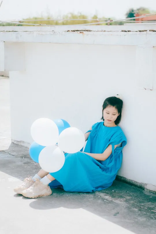 一个小女孩穿着蓝色裙子 靠在白色墙上拿着气球。