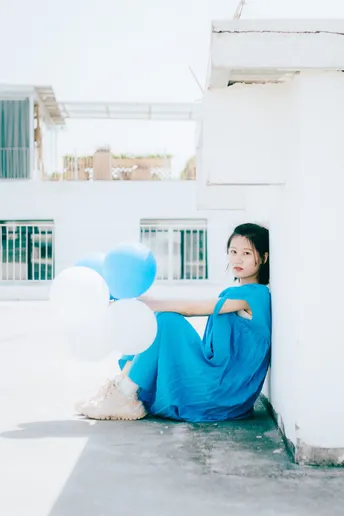 一个穿着蓝色连衣裙的年轻女子坐在一个白色建筑物的地板上 手里拿着气球。
