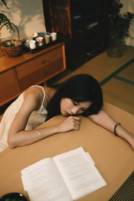 一个年轻女子躺在地上 倚靠在一张桌子旁边 桌子上放着一本打开的书。