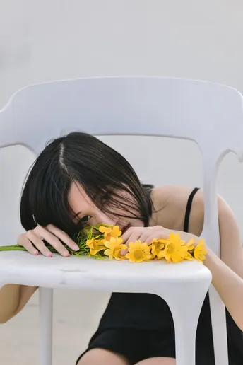 一个穿着黑色连衣裙的年轻女子坐在白色椅子上 用黄色花朵遮住她的脸。
