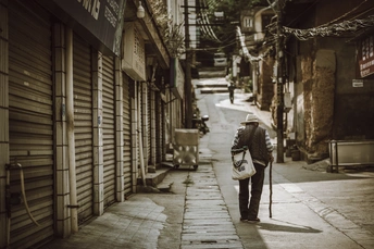 一个老人背着书包 手拿拐杖 走在建筑物之间的狭窄小巷。