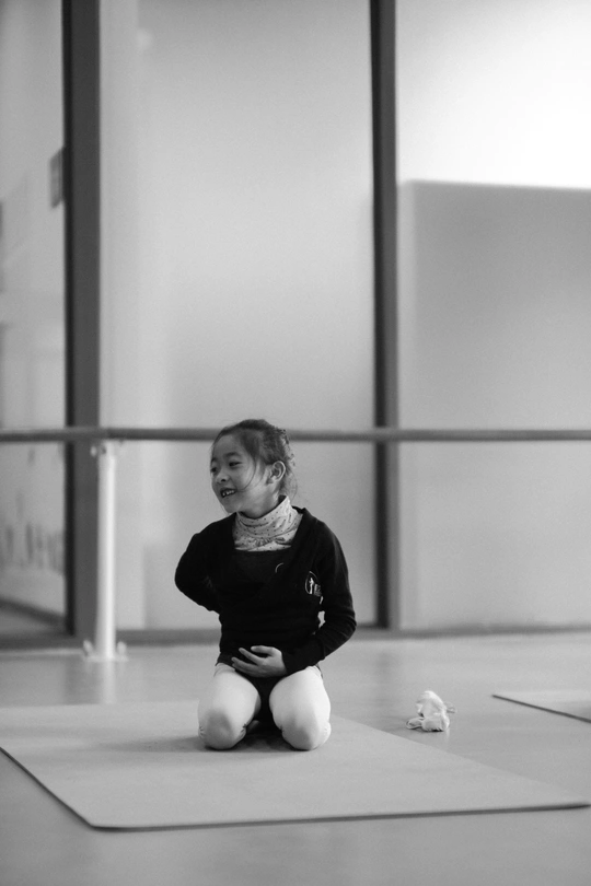 一个小女孩坐在瑜伽室的地垫上 微笑着 后面有一个小孩子在地上坐着。