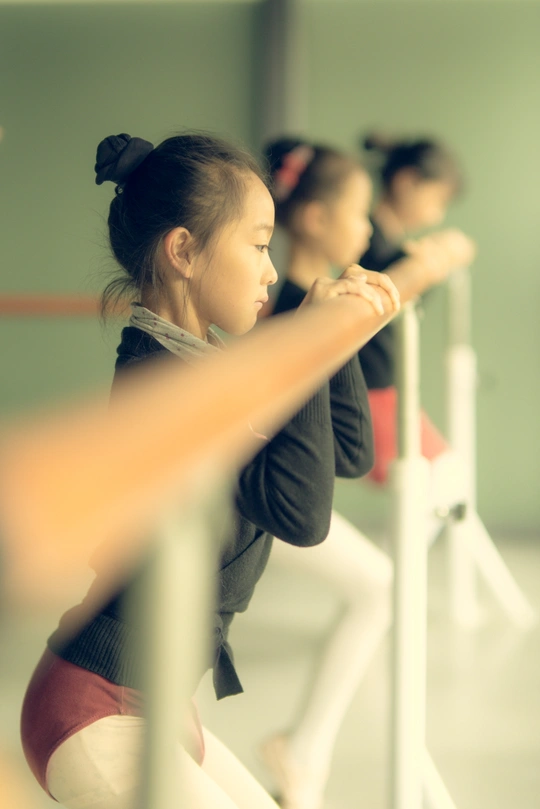 一个小女孩站在镜子前练习芭蕾舞 其他年轻舞者观看。