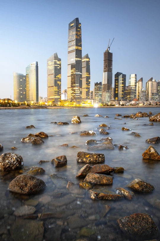 河岸上有岩石 前景是城市天际线上的摩天大楼 前景中还有水域。