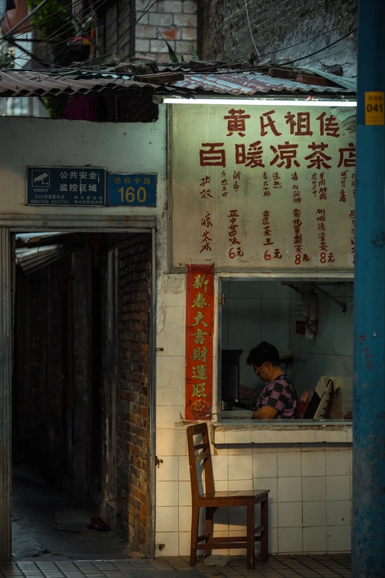 夜晚的一个小巷子里 有一个小建筑 上面写着“面馆” 一个妇女坐在餐馆前面的一张桌子旁。