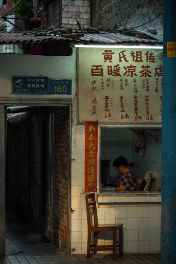 夜晚的一个小巷子里 有一个小建筑 上面写着“面馆” 一个妇女坐在餐馆前面的一张桌子旁。