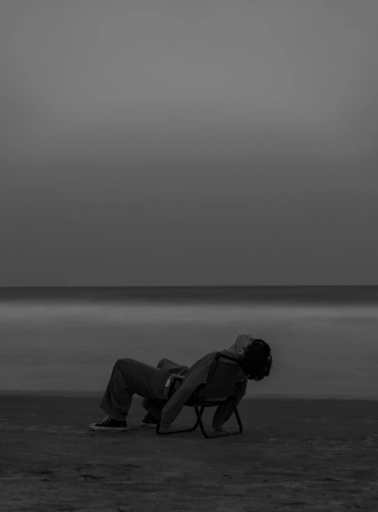 一个人坐在沙滩上的椅子上 手里拿着一把伞 背景中有一个面对水域坐着的男人。