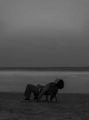 一个人坐在沙滩上的椅子上 手里拿着一把伞 背景中有一个面对水域坐着的男人。