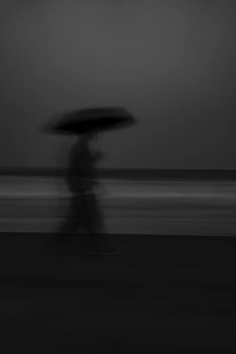 一张黑白图像 描绘一个人在夜晚的沙滩上持伞行走