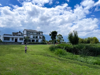 一个人站在草地上 天空中有一只风筝 背景有一座大型建筑。