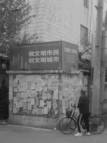 黑白照片中 一位妇女骑着自行车走在一条街道上 街道前方有一面被各种标志覆盖的墙和一座建筑。