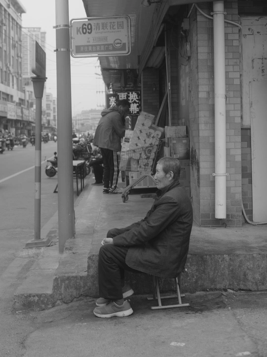 一张黑白照片 一位老人坐在人行道上的一把椅子里。