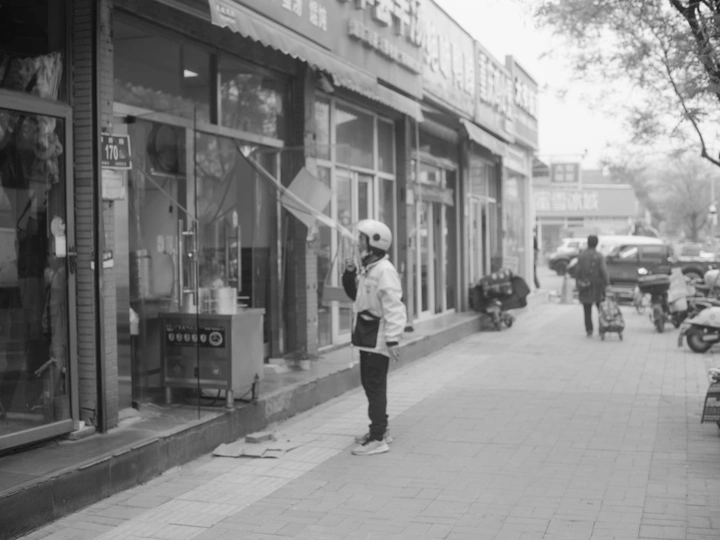 一张黑白照片 一个戴头盔的年轻男子在城市的街道上滑板 街道上有商店。