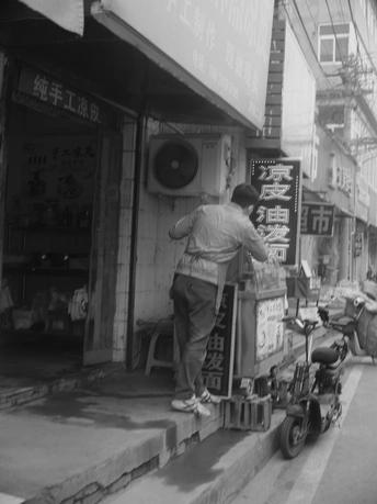 一张黑白照片 一个站在人行道上 摩托车前面的商店门口的男子。