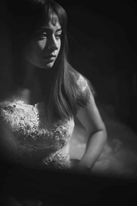 一个黑白肖像画 描绘了一位穿着白色连衣裙的年轻女子 坐在一个黑暗房间的镜子前。