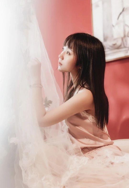 一个穿着婚纱带着面纱的年轻女子坐在床上 凝视着窗外的一扇红色窗户。