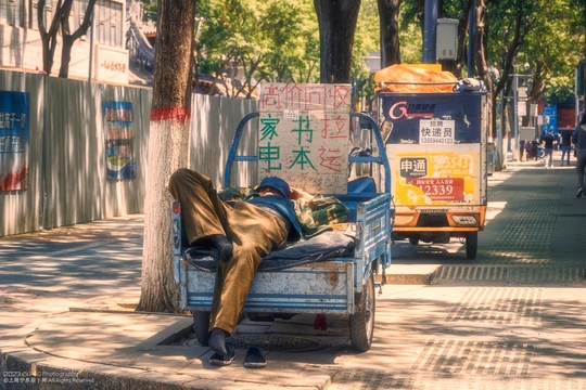 一个男子躺在床上 倚靠在树下的长凳上 在繁忙街道的人行道上休息。
