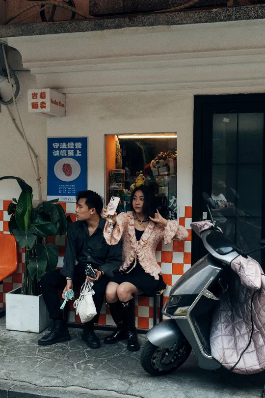 一个人坐在摩托车上 一个女人坐在建筑物前面的长椅上 用手机拍照。