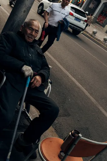 一位老人坐在人行道旁的一把椅子上 旁边是一条繁忙的街道。