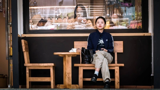 一位年轻男子坐在一家餐厅前的木凳上 窗外坐着一个人。