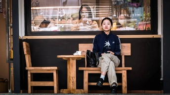 一位年轻男子坐在一家餐厅前的木凳上 窗外坐着一个人。