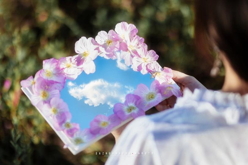 一位女子手持一面镜子 背景是粉红色花朵和蓝天。