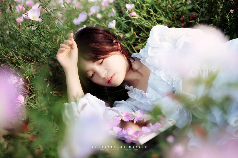 一位穿着白色连衣裙的年轻女子躺在紫花丛中 手里拿着一本书。