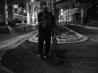 一张黑白照片 一个背书包的男人站在夜晚人行道上 旁边有一辆自行车。