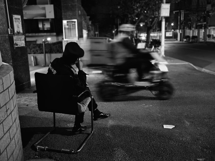 一张黑白照片 一个人坐在街边的一把椅子上 晚上 人们骑摩托车。