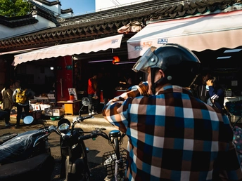 戴着头盔的男子站在一辆自行车旁边 市场前有人们在走动。