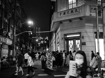 一张黑白照片 描绘了一个夜晚在拥挤的城市街道人行道上阅读报纸的女人。