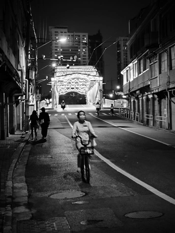一个人在夜晚骑自行车穿过城市街道 背景中有桥和其他人。