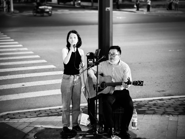 一个黑白镜头 展示一个男子在弹吉他 一个女子站在人行道上 两人都在演奏音乐。