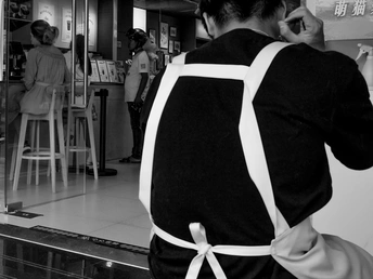 一个穿黑白格 apron 的女人正在用手机讲话 一个男子站在柜台后面。