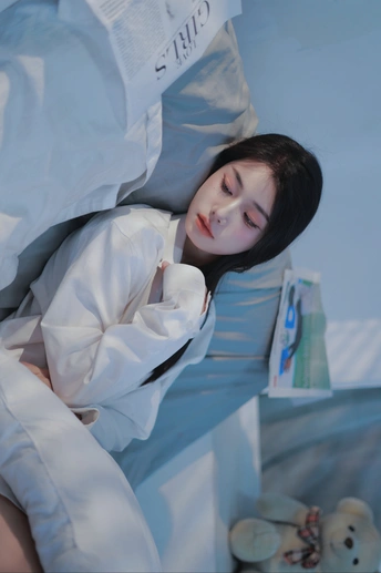 一个年轻女子躺在病床上 床单铺开 枕头上躺着一个泰迪熊。