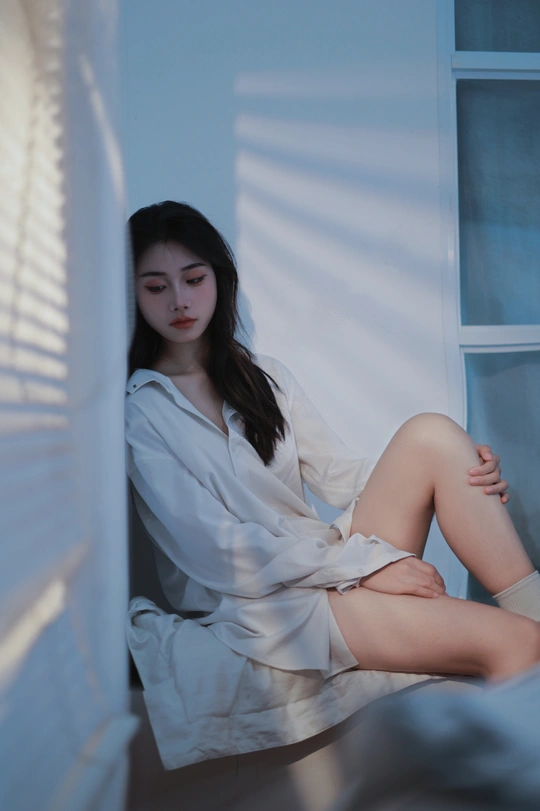 一个年轻女子穿着白色衬衫坐在床上 凝视着窗户。