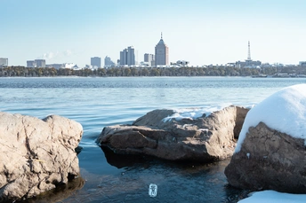 湖面结冰 岩石上有雪和冰 背景中有一座城市。